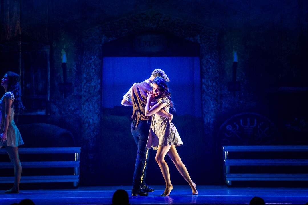 A male and a female dancer. The female dancer is leaning into the male dancer, who has his hand around her waist. The lights are dark blue, but the dancers are still well illuminated.