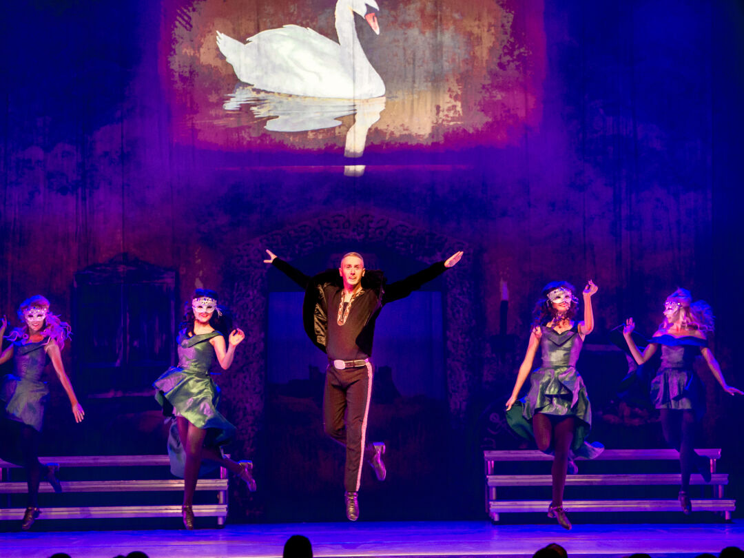 several dancers are lined up on stage. Male dancer stands in the middle, mid jump. Two female dancers stand either side of him.