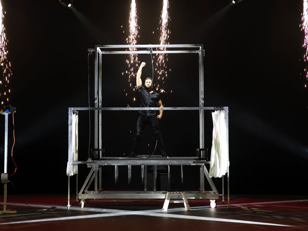 Promotional image for Spellbound. Performer stands in a metal cage with his right arm up, showcasing his magic tricks. Sparks come down from the ceiling above him.