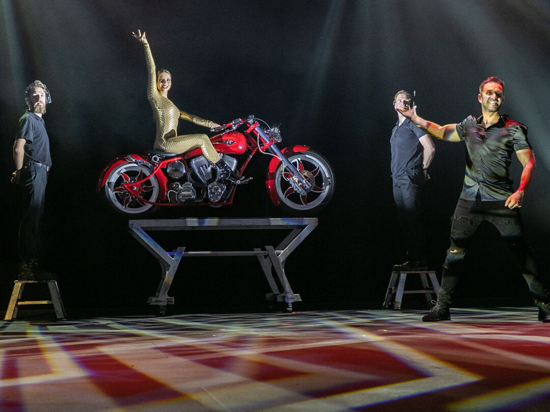 Female performer sits on a red motorbike with her right hand in the air, with two male volunteers standing either side of her. Male performer stands in front of her, gesturing to her.