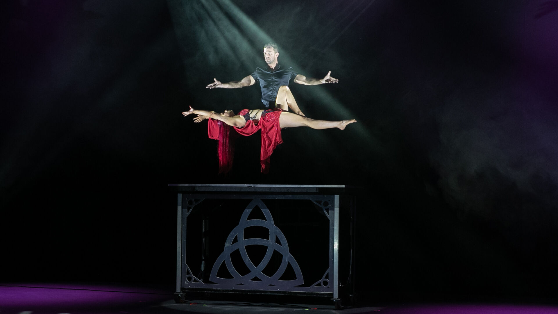 Male performer stands over female performer, performing levitating magic trick. Female performer is suspended over a dark box with silver accents, seeming to float in the air.