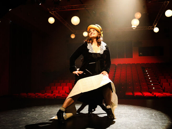 Performer Susanne Campbell is sitting on a chair on the stage with a spotlight